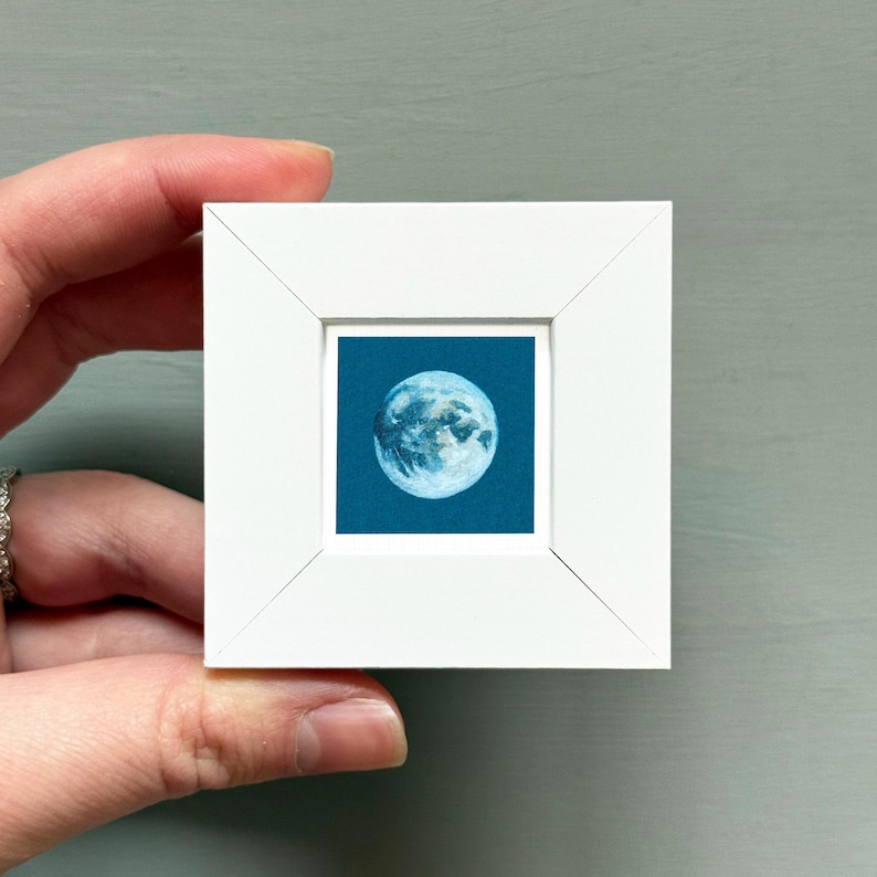 A hand is holding a small white picture frame containing a depiction of the moon.