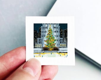 May include: A small square print featuring a painting of a brightly lit Christmas tree standing in front of a tall gray building at night. The scene includes a light-colored ground with small figures of people scattered around the base of the tree.