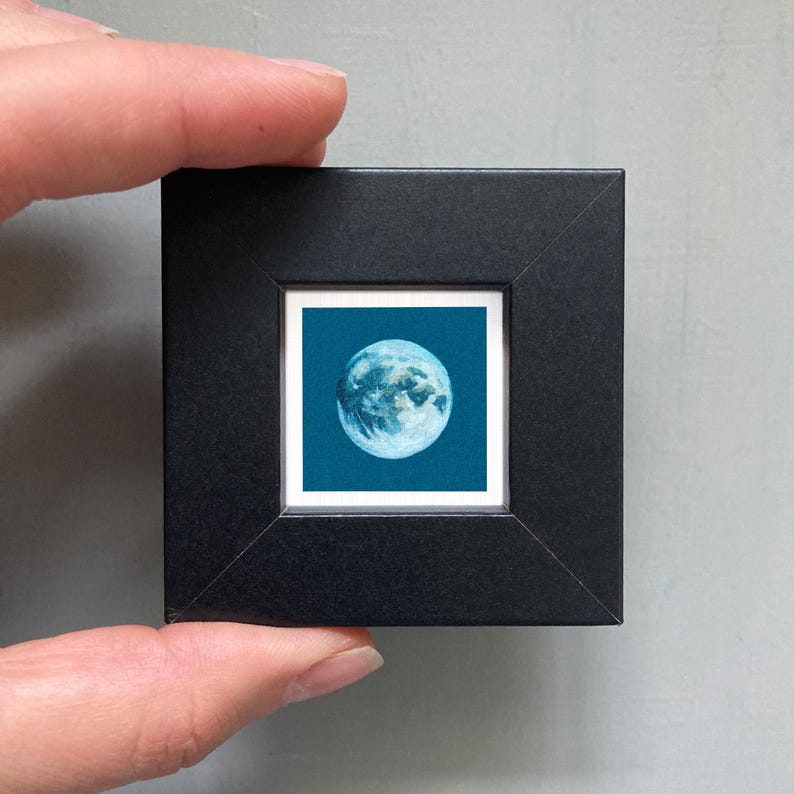 A person's hand is holding a small black square frame containing a framed image of a moon.