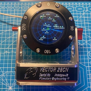May include: A circular black radar display with a blue screen showing flight data, mounted on a silver metal stand. The base features the text VECTOR 28CN, Serial No Prototype-02, and Planetary Engineering, with a small fish logo graphic.