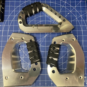 Puede incluir: Tres mangos de metal de color plateado con forma triangular o curva, cada uno con una sección envuelta en cordón negro. Las superficies metálicas son lisas y reflectantes, con múltiples orificios circulares preperforados en los bordes.