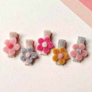 May include: Five flower-shaped hair clips in shades of pink, grey, fuchsia, yellow, and pink with grey centres. Each clip is attached to a neutral-toned fabric base. The background is white with a diagonal pink and white striped pattern.