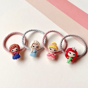 May include: Four colorful hair ties with princess character charms. The charms are of Ariel, Cinderella, Elsa, and Rapunzel. The hair ties are pink and gray.