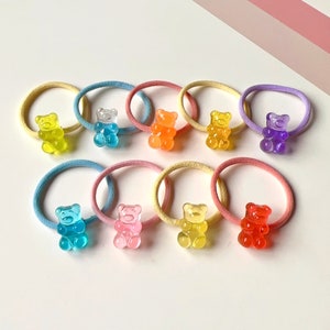 Può includere: Assortimento di elastici per capelli a forma di orsacchiotto gommoso colorati. Ogni elastico per capelli presenta un ciondolo a forma di orsacchiotto gommoso trasparente di un colore diverso, tra cui giallo, arancione, rosa, blu, viola e rosso.