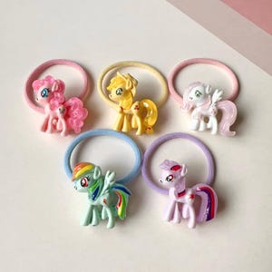 May include: Set of five colorful hair ties with My Little Pony characters. Each tie features a different pony with a unique color scheme and design. The ponies are pink, yellow, white, green, and purple.
