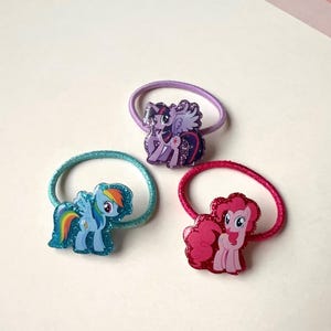 May include: Three colorful hair ties with My Little Pony characters. The hair ties are purple, blue, and pink. Each hair tie features a different pony character: Twilight Sparkle, Rainbow Dash, and Pinkie Pie.