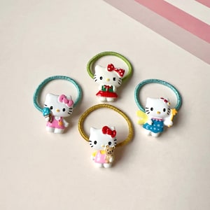 May include: Four colourful Hello Kitty hair ties with different designs. Each tie features a Hello Kitty character with a different outfit and accessory. The hair ties are made of elastic and have a glitter finish.