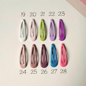 May include: A collection of ten oval-shaped metal snap hair clips arranged in two rows of five. Each clip features a smooth, solid-colored enamel finish in shades of white, pink, green, lavender, purple, peach, mauve, plum, blue, and hot pink.