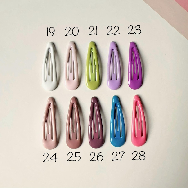 May include: A collection of ten oval-shaped metal snap hair clips arranged in two rows of five. Each clip features a smooth, solid-colored enamel finish in shades of white, pink, green, lavender, purple, peach, mauve, plum, blue, and hot pink.