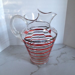 May include: A clear glass pitcher with red and silver stripes. The pitcher has a wide mouth and a handle.