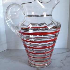 May include: A clear glass pitcher with a red and silver striped design. The pitcher has a wide mouth and a handle.