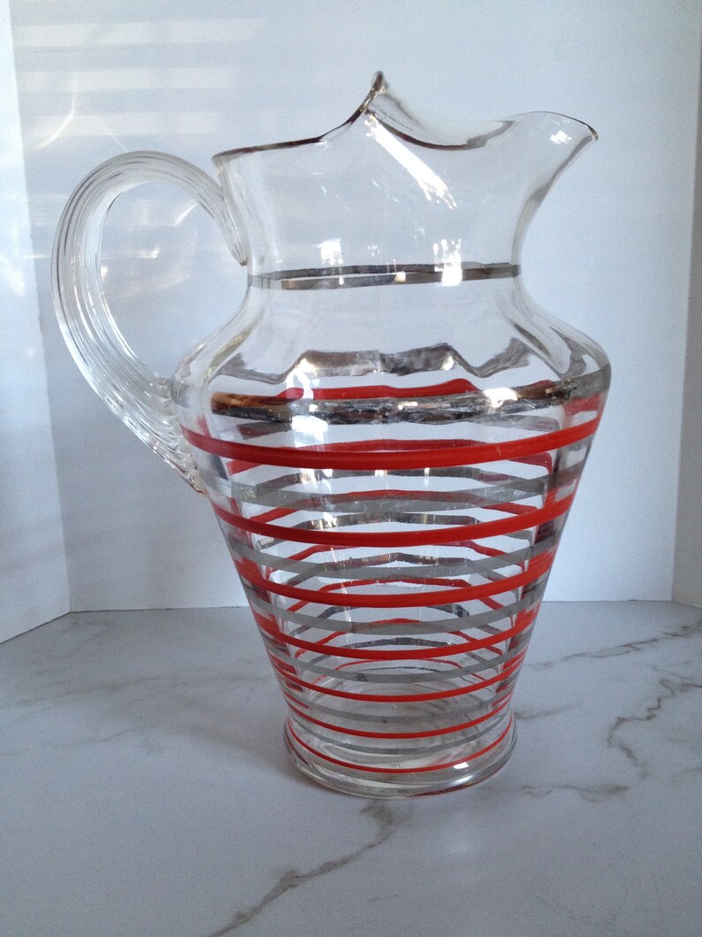 May include: A clear glass pitcher with a red and silver striped design. The pitcher has a wide mouth and a handle.