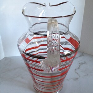 May include: A clear glass pitcher with a red and gray striped design. The pitcher has a wide mouth and a handle.