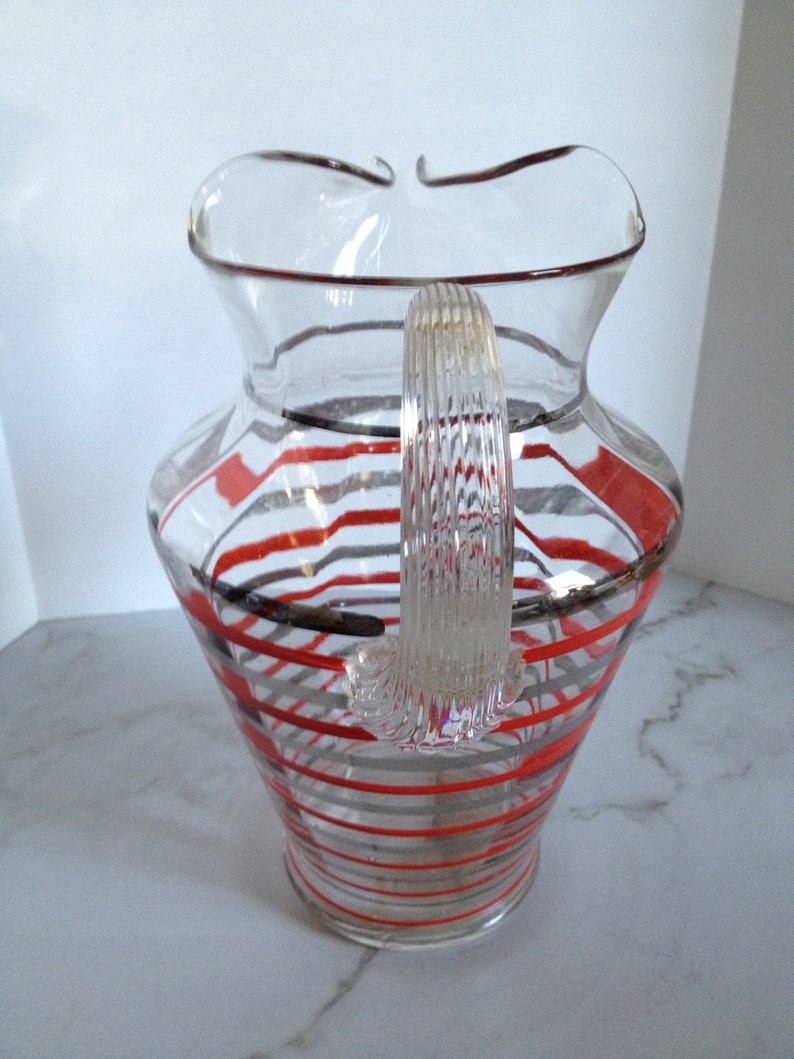 May include: A clear glass pitcher with a red and gray striped design. The pitcher has a wide mouth and a handle.