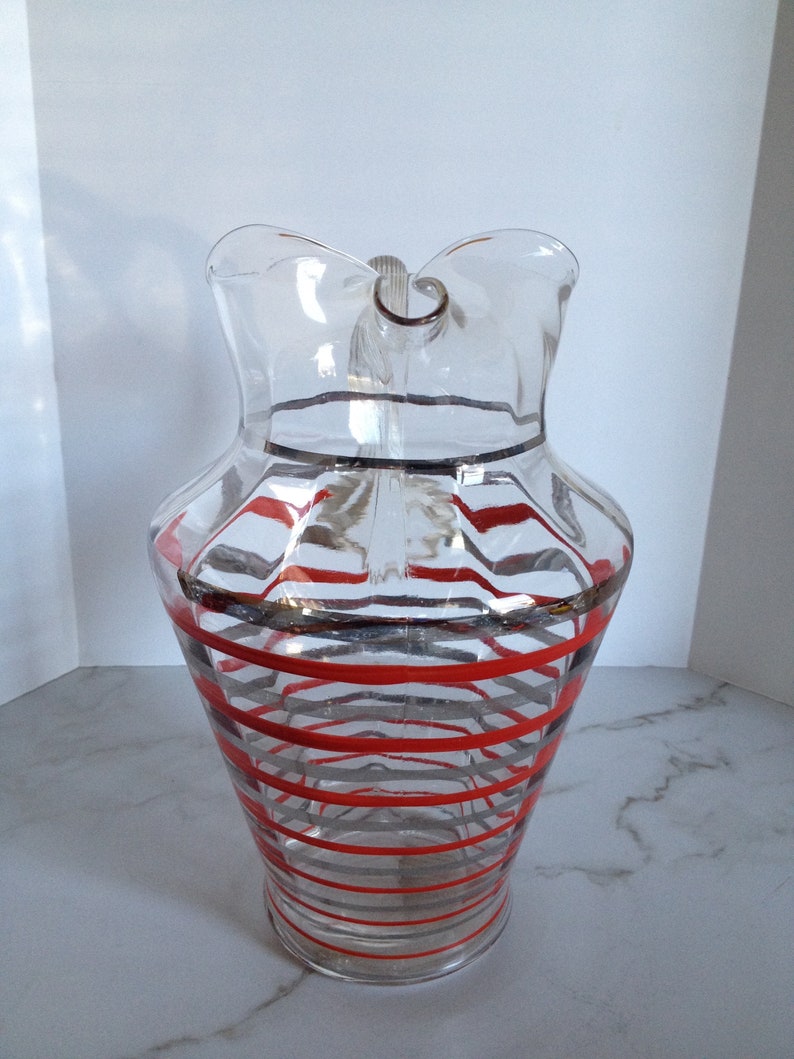 May include: A clear glass pitcher with a red and gray striped design. The pitcher has a wide mouth and a handle.
