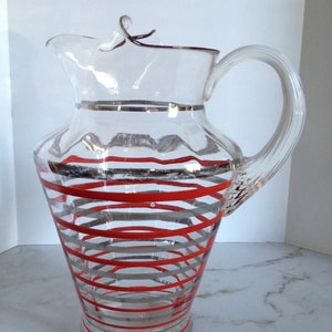 May include: A clear glass pitcher with a red and silver striped design. The pitcher has a handle and a spout.
