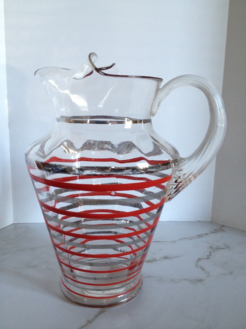 May include: A clear glass pitcher with a red and silver striped design. The pitcher has a handle and a spout.