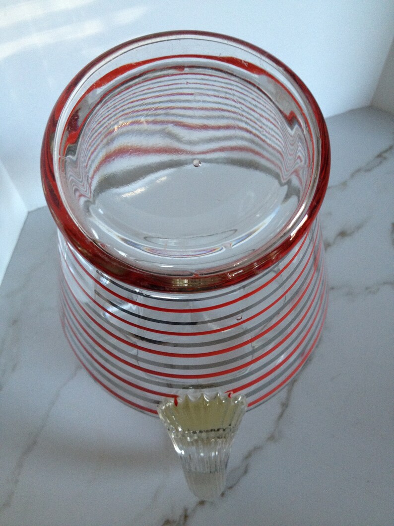 May include: A clear glass pitcher with a red and gray striped pattern. The pitcher has a handle with a decorative design.