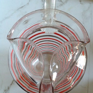 May include: A clear glass pitcher with a red and gray striped design. The pitcher has a handle and a wide mouth.