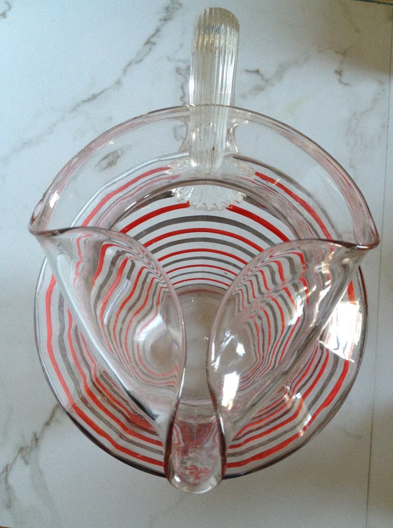 May include: A clear glass pitcher with a red and gray striped design. The pitcher has a handle and a wide mouth.