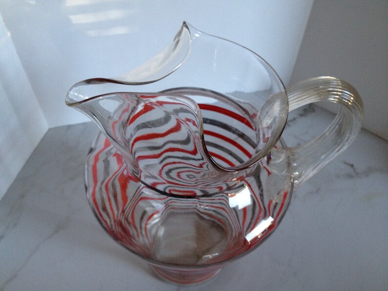 May include: A clear glass pitcher with a red and gray striped design. The pitcher has a wide mouth and a curved handle.