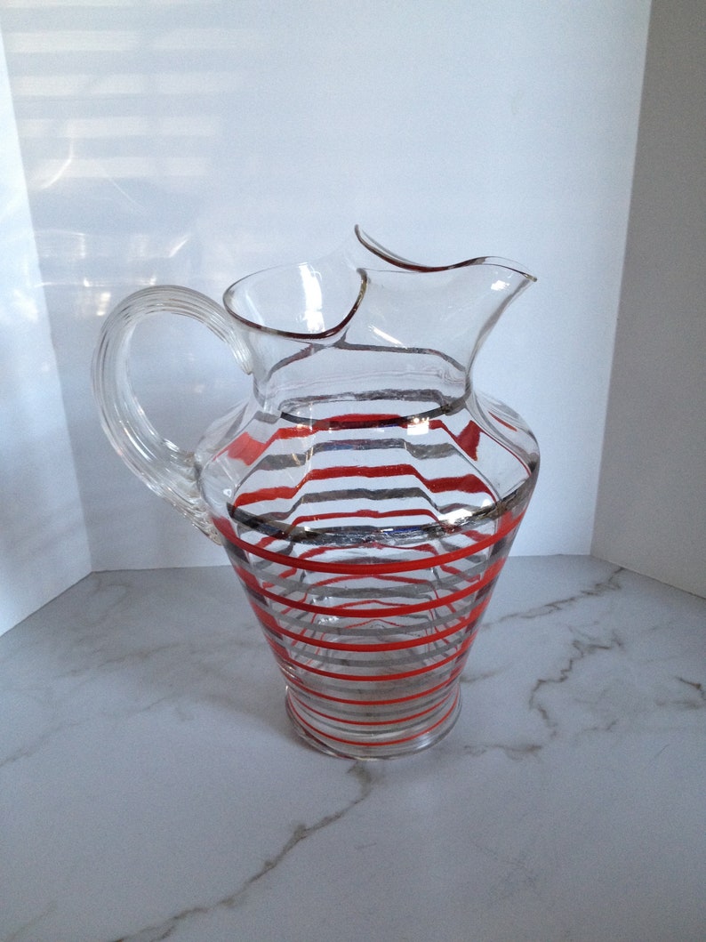 May include: A clear glass pitcher with red and silver stripes. The pitcher has a wide mouth and a handle.