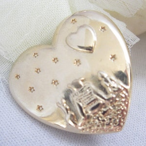 May include: A gold-colored heart-shaped pendant with a small heart and star accents. The pendant features a detailed relief design of a bell and other elements. The pendant is likely made of metal.