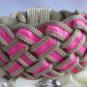 Puede incluir: Un brazalete tejido rosa y dorado con un diseño trenzado. El brazalete está hecho de cuero y metal.
