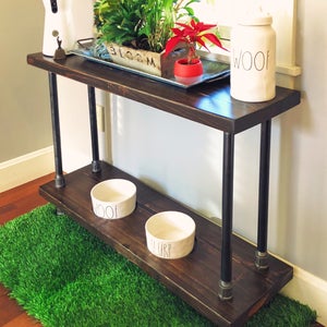 May include: A dark brown wooden console table with black metal pipe legs. The table has a top shelf and a lower shelf. Two white ceramic dog bowls are on the lower shelf. A green artificial grass mat is in front of the table. The table has a white ceramic jar with the word "WOOF" on it.