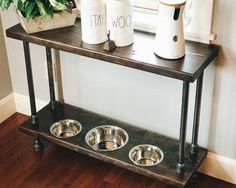 May include: A dark brown wooden table with black metal legs. The table has two shelves. The top shelf has two white ceramic canisters with the words "STAY" and "WOO" on them. The bottom shelf has three stainless steel dog bowls.