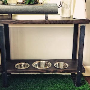 May include: A dark brown wooden table with black metal legs and a lower shelf. Three stainless steel dog bowls are on the shelf. The table is in front of a green artificial grass rug.