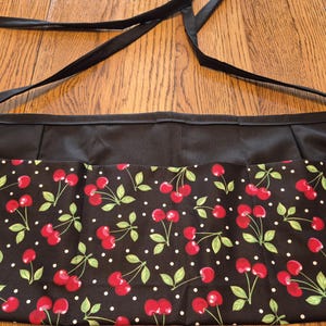 May include: Black apron with a cherry pattern. The apron features a black satin waistband and ties. The lower portion of the apron is decorated with red cherries, green leaves, and white polka dots on a black background.