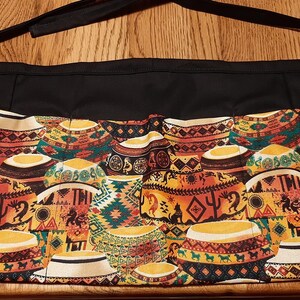 May include: Black apron with multiple pockets and a colorful patterned bottom featuring a repeating design of yellow, orange, red, green, and blue geometric shapes. The pattern resembles traditional African pottery.