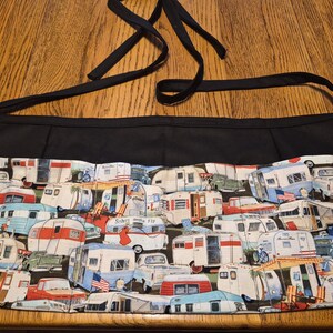 May include: A black waist apron with a colorful pattern of vintage travel trailers. The apron has a black waistband and ties, and the fabric features various trailer designs in red, blue, and white.