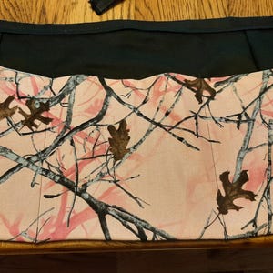 May include: A black and pink camouflage apron with a TrueTimber logo. The apron features a pink and white camouflage pattern with brown leaves and black branches. The top of the apron is solid black.
