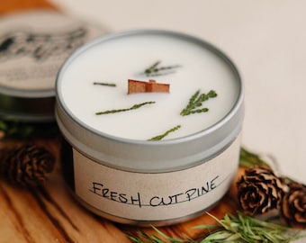 May include: A silver tin candle with a white wax top and a label that reads "Fresh Cut Pine". The candle is surrounded by pine needles and pine cones on a wooden surface.