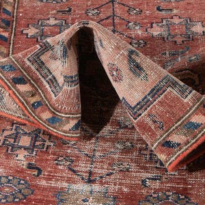 May include: A rectangular area rug featuring a faded terracotta red base with intricate geometric and floral patterns in shades of blue, cream, and dark brown. The rug is folded over in the center and includes a white tag labeled No: 5829.