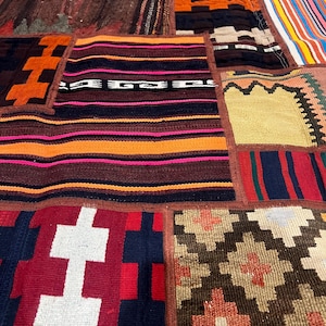 May include: A patchwork rug composed of various rectangular textile segments featuring diverse woven patterns and colors including deep red, orange, dark brown, black, white, and blue. The sections are joined by visible brown stitched seams.