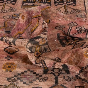 May include: A woven rug featuring a faded dusty rose background with intricate geometric patterns in black, mustard yellow, and muted blue. The textile displays a repeating motif of stylized floral shapes and diamonds across the surface.