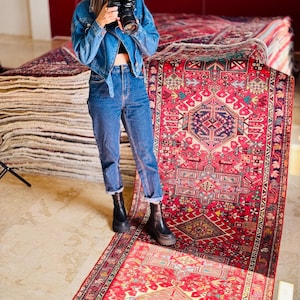 May include: A vibrant red Persian rug with detailed geometric designs is shown on a light floor. The rug has a central motif and borders with multiple colours. Other rugs are stacked in the background.