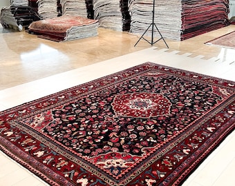 May include: A rectangular area rug featuring a dark black central field with a dense floral pattern in shades of red, blue, and cream. The rug has a wide red border with intricate geometric and botanical motifs and a fringed edge on the ends.