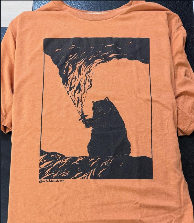 May include: A burnt orange short-sleeved t-shirt featuring a black graphic print of a cat holding a sword with flames rising from the blade. The design is enclosed in a rectangular border and includes the text @artofdonialryan at the bottom.