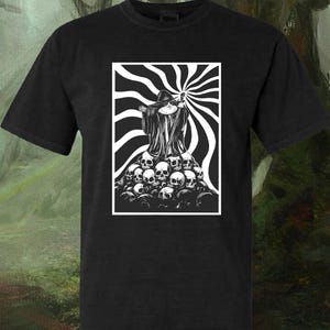 Könnte beinhalten: Ein schwarzes Kurzarm-T-Shirt mit einem weißen Grafikdruck einer in eine Robe gekleideten Figur mit spitzem Hut, die auf einem Haufen Totenköpfe steht. Der Hintergrund der Grafik besteht aus strahlenden schwarz-weißen Wellenlinien in einem Rahmen.