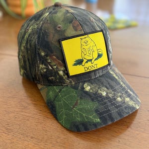Puede incluir: Una gorra de béisbol con estampado de camuflaje y un parche rectangular amarillo en la parte delantera. El parche muestra un dibujo lineal negro de un gato sentado sobre hierba verde con la palabra DONT impresa en letras mayúsculas negras.