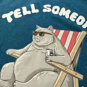 May include: Teal t-shirt featuring a cartoon cat wearing sunglasses, relaxing in a beach chair, and holding a beverage. The text "TELL SOMEONE" is arched across the top in white letters. The cat is gray, and the chair has red and white stripes.
