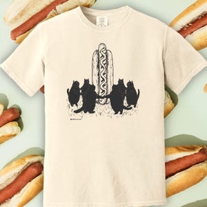 May include: A cream-colored short-sleeve t-shirt featuring a black ink illustration of five black cats standing in a circle holding hands around a large hot dog. The graphic is centered on the chest and includes small text below the cats.