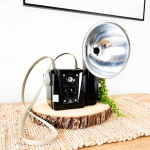 May include: A vintage black and silver camera with a flash attachment, sitting on a wooden base with a natural fiber rug underneath.