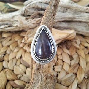 May include: A teardrop-shaped ring with a large, polished, purple gemstone set in a silver-colored metal. The ring has a braided rope design around the gemstone and sits on a piece of driftwood.