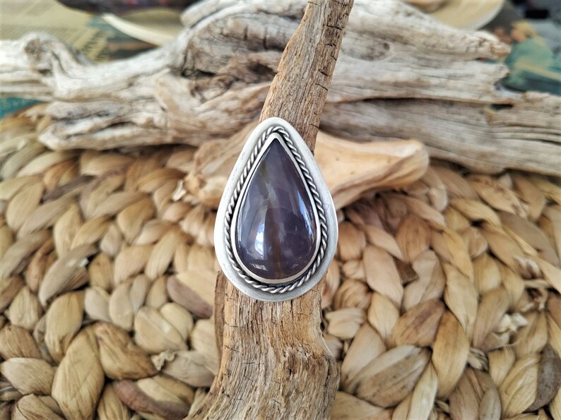 May include: A teardrop-shaped ring with a large, polished, purple gemstone set in a silver-colored metal. The ring has a braided rope design around the gemstone and sits on a piece of driftwood.