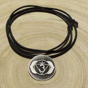 Op de afbeelding: Een zilverkleurige hangerketting met een chakra-symbool, hangend aan een zwart koord. De hanger heeft een ronde vorm met een gedetailleerd ontwerp. De ketting wordt getoond op een lichtbruin houten oppervlak.